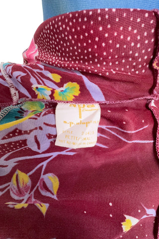 Vintage 1970s dress in burgundy red with a tulip and dianthus floral print. Features short flutter sleeves and a round neckline that can be gathered with the bow tie. The bodice closes with three white diamond shaped plastic buttons. The neckline and sleeves are finished with tiny white scalloped eyelet trim.