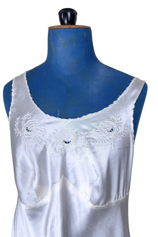 Vintage 1940s nightgown or slip, with cutwork embroidery across the bust, a pointed midriff yoke with vertical bust darts, and scalloped edging around the wide round neckline and hem. The hem has a small kick slit at the left, accented with floral appliqué and embroidery.