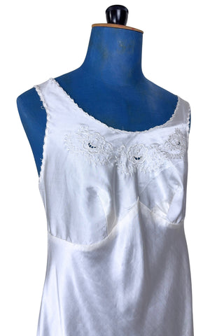 Vintage 1940s nightgown or slip, with cutwork embroidery across the bust, a pointed midriff yoke with vertical bust darts, and scalloped edging around the wide round neckline and hem. The hem has a small kick slit at the left, accented with floral appliqué and embroidery.