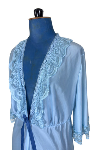 Vintage 1970s negligee robe in sky blue with wide lace trim along the deep V neckline and three-quarter length sleeve cuffs. Finished with a decorative royal blue bow. Closes at the waist with a single button.