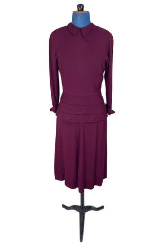 Vintage 1930s day dress in burgundy wool crepe, featuring a small pointed collar, long sleeves with turned cuffs, and three wide bias-cut tucks at the hip forming a peplum flounce around the front. In the back, the peplum becomes a waterfall pleat panel down the middle back. The dress closes in the back with a metal zipper, and is unlined. 