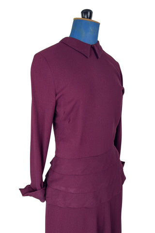 Vintage 1930s day dress in burgundy wool crepe, featuring a small pointed collar, long sleeves with turned cuffs, and three wide bias-cut tucks at the hip forming a peplum flounce around the front. In the back, the peplum becomes a waterfall pleat panel down the middle back. The dress closes in the back with a metal zipper, and is unlined. 