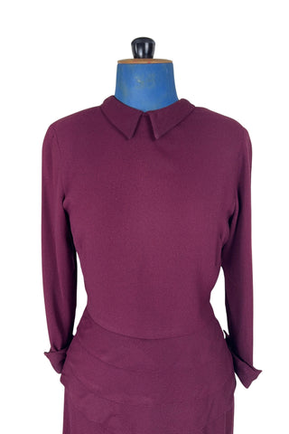 Vintage 1930s day dress in burgundy wool crepe, featuring a small pointed collar, long sleeves with turned cuffs, and three wide bias-cut tucks at the hip forming a peplum flounce around the front. In the back, the peplum becomes a waterfall pleat panel down the middle back. The dress closes in the back with a metal zipper, and is unlined. 