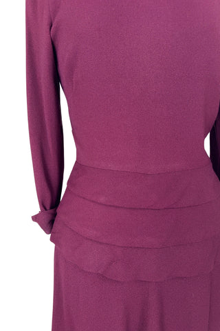 Vintage 1930s day dress in burgundy wool crepe, featuring a small pointed collar, long sleeves with turned cuffs, and three wide bias-cut tucks at the hip forming a peplum flounce around the front. In the back, the peplum becomes a waterfall pleat panel down the middle back. The dress closes in the back with a metal zipper, and is unlined. 