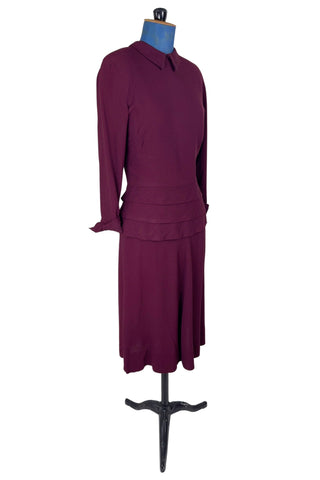 Vintage 1930s day dress in burgundy wool crepe, featuring a small pointed collar, long sleeves with turned cuffs, and three wide bias-cut tucks at the hip forming a peplum flounce around the front. In the back, the peplum becomes a waterfall pleat panel down the middle back. The dress closes in the back with a metal zipper, and is unlined. 