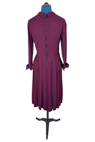 Vintage 1930s day dress in burgundy wool crepe, featuring a small pointed collar, long sleeves with turned cuffs, and three wide bias-cut tucks at the hip forming a peplum flounce around the front. In the back, the peplum becomes a waterfall pleat panel down the middle back. The dress closes in the back with a metal zipper, and is unlined. 