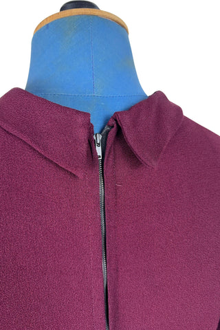 Vintage 1930s day dress in burgundy wool crepe, featuring a small pointed collar, long sleeves with turned cuffs, and three wide bias-cut tucks at the hip forming a peplum flounce around the front. In the back, the peplum becomes a waterfall pleat panel down the middle back. The dress closes in the back with a metal zipper, and is unlined. 