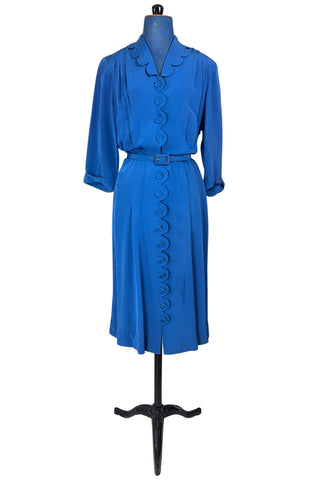 Vintage 1940s day dress in royal blue rayon with scalloped collar and front button placket, which closes with 14 functional self-fabric buttons down the front. The dress has three quarter sleeves with folded cuffs, and a matching self-fabric belt with a rectangular covered buckle. 
Condition Note: The dress is in very good vintage condition, with an inconspicuous small darkened area on the lower back of the skirt about 1-2” long.