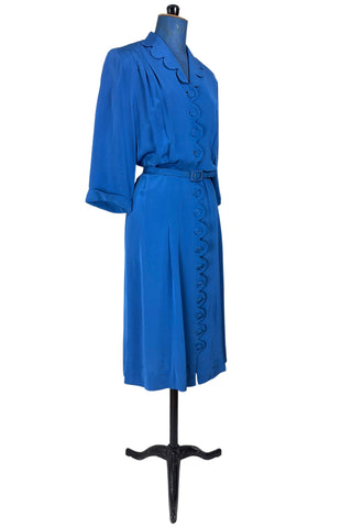 Vintage 1940s day dress in royal blue rayon with scalloped collar and front button placket, which closes with 14 functional self-fabric buttons down the front. The dress has three quarter sleeves with folded cuffs, and a matching self-fabric belt with a rectangular covered buckle. 
Condition Note: The dress is in very good vintage condition, with an inconspicuous small darkened area on the lower back of the skirt about 1-2” long.