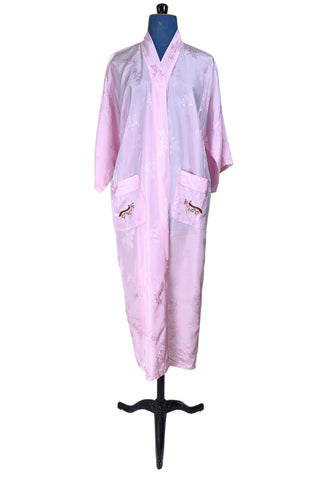 Vintage 1960s kimono style robe in light pink rayon damask, with embroidered crane birds and nature scenes. Features a deep V neckline, three-quarter length wide sleeves, and two large pockets decorated with embroidery. The back of the robe has a large embroidered motif.