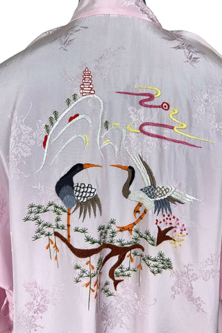 Vintage 1960s kimono style robe in light pink rayon damask, with embroidered crane birds and nature scenes. Features a deep V neckline, three-quarter length wide sleeves, and two large pockets decorated with embroidery. The back of the robe has a large embroidered motif.
