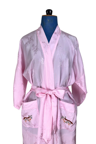 Vintage 1960s kimono style robe in light pink rayon damask, with embroidered crane birds and nature scenes. Features a deep V neckline, three-quarter length wide sleeves, and two large pockets decorated with embroidery. The back of the robe has a large embroidered motif.