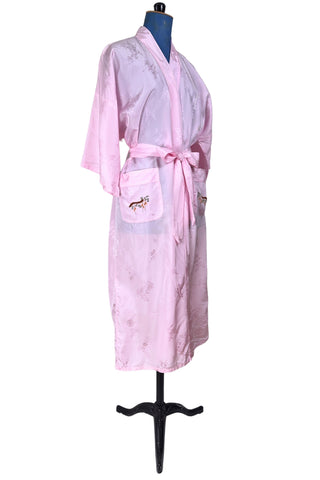 Vintage 1960s kimono style robe in light pink rayon damask, with embroidered crane birds and nature scenes. Features a deep V neckline, three-quarter length wide sleeves, and two large pockets decorated with embroidery. The back of the robe has a large embroidered motif.