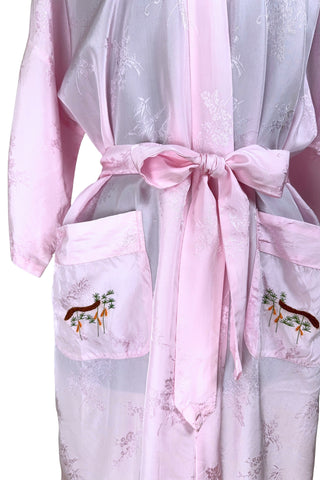 Vintage 1960s kimono style robe in light pink rayon damask, with embroidered crane birds and nature scenes. Features a deep V neckline, three-quarter length wide sleeves, and two large pockets decorated with embroidery. The back of the robe has a large embroidered motif.