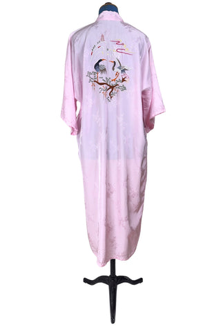 Vintage 1960s kimono style robe in light pink rayon damask, with embroidered crane birds and nature scenes. Features a deep V neckline, three-quarter length wide sleeves, and two large pockets decorated with embroidery. The back of the robe has a large embroidered motif.