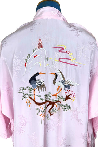 Vintage 1960s kimono style robe in light pink rayon damask, with embroidered crane birds and nature scenes. Features a deep V neckline, three-quarter length wide sleeves, and two large pockets decorated with embroidery. The back of the robe has a large embroidered motif.