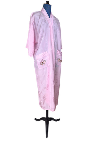 Vintage 1960s kimono style robe in light pink rayon damask, with embroidered crane birds and nature scenes. Features a deep V neckline, three-quarter length wide sleeves, and two large pockets decorated with embroidery. The back of the robe has a large embroidered motif.