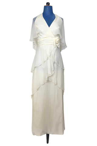 Delicate and lovely 1970s gown in layered cream white silk chiffon. This heavenly dress is composed of layers of cream silk chiffon in an Edwardian-inspired column silhouette. Featuring a deep V halter neckline, the bodice has a built-in cape of sheer chiffon which covers the shoulders and back to the waist. A gathered sash of chiffon is set-off with a large rosette to one side. The sash fastens inconspicuously under the flower so that the fasteners are hidden. The back of the neck has different loop...