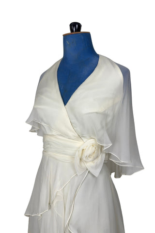 Delicate and lovely 1970s gown in layered cream white silk chiffon. This heavenly dress is composed of layers of cream silk chiffon in an Edwardian-inspired column silhouette. Featuring a deep V halter neckline, the bodice has a built-in cape of sheer chiffon which covers the shoulders and back to the waist. A gathered sash of chiffon is set-off with a large rosette to one side. The sash fastens inconspicuously under the flower so that the fasteners are hidden. The back of the neck has different loop...