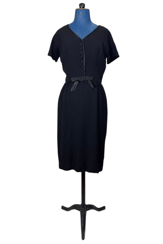 Vintage 1950s black cocktail dress with a large black satin bow at the waist. The dress features a V neckline trimmed with satin, with tiny covered satin buttons down the bodice. The waist is accentuated with small black satin piping.

Designed with a classic 1950s hourglass silhouette, the dress has a nipped waist, and hugs the wearer’s curves through the bust and hips. This style is often described as a “wiggle dress”.

The dress closes in the back with a Clix metal zipper, and is unlined.