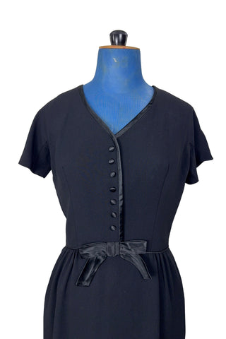 Vintage 1950s black cocktail dress with a large black satin bow at the waist. The dress features a V neckline trimmed with satin, with tiny covered satin buttons down the bodice. The waist is accentuated with small black satin piping.

Designed with a classic 1950s hourglass silhouette, the dress has a nipped waist, and hugs the wearer’s curves through the bust and hips. This style is often described as a “wiggle dress”.

The dress closes in the back with a Clix metal zipper, and is unlined.