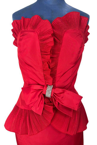 Vintage late 1980s or early 1990s cocktail dress in bold red acetate faille fabric with subtle ribbed texture. The strapless bodice has a wrapped appearance, trimmed with wide panels of accordion pleated ruffles. A large bow at the waist is crowned with a wide band of rhinestones.

Designed with an hourglass silhouette, the dress hugs and accentuates curves for a bombshell fit. 

Marked size 9/10 (vintage sizing - see measurements section at right).

Designed by Collections JS. Made in Canada.
